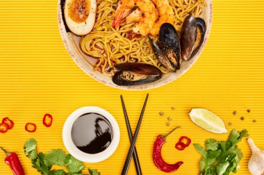 Taze malzemelerin yanında deniz mahsulleri ramen ve sarı yüzeyde yemek çubukları.
