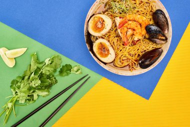 Taze malzemelerin yanında baharatlı deniz mahsulü ramen ve çok renkli yüzey üzerinde çubuklar.