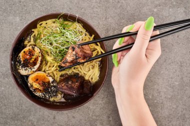 cropped view of woman holding chopsticks near spicy meat ramen on grey surface clipart