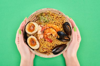 Baharatlı deniz mahsullü ramen ile yeşil yüzeyde elinde kase tutan bir kadın görüntüsü.