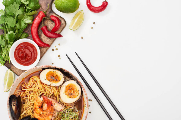 top view of spicy seafood ramen near fresh ingredients and chopsticks on white surface