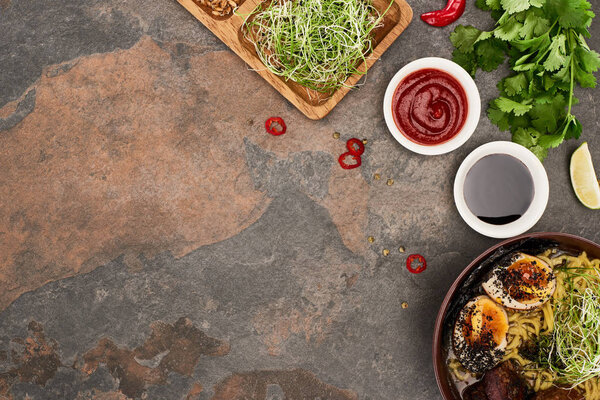 top view of spicy meat ramen near fresh ingredients and sauces on stone surface