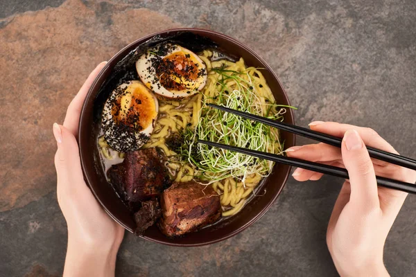 cropped view of woman eating spicy meat ramen with chopsticks on stone surface