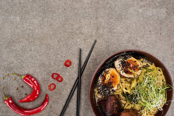 top view of spicy meat ramen near chili pepper and chopsticks on grey concrete surface