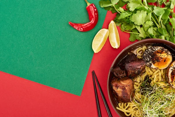 在绿色和红色表面的新鲜配料和筷子附近的辣肉类拉面的顶部视图 — 图库照片