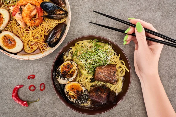 Cropped view of woman holding chopsticks near spicy seafood and meat ramen on grey surface — Stock Image Cropped View Woman Holding Chopsticks Spicy Seafood Meat Ramen Grey Stock Image