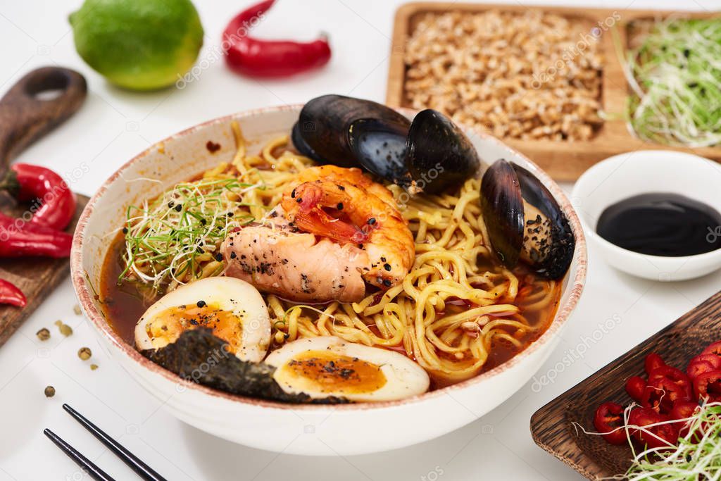 spicy seafood ramen near fresh ingredients and chopsticks on white surface