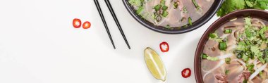 top view of pho in bowl near chopsticks, lime, chili and soy sauces and coriander on white background, panoramic shot clipart