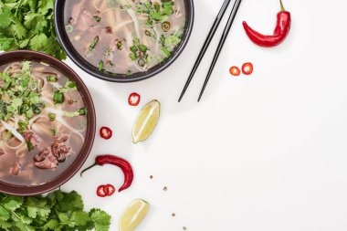 top view of pho in bowls near chopsticks, lime, chili and coriander on white background clipart