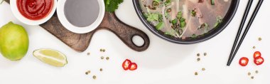 top view of pho in bowl near chopsticks, lime, chili and soy sauces and coriander on white background, panoramic shot clipart