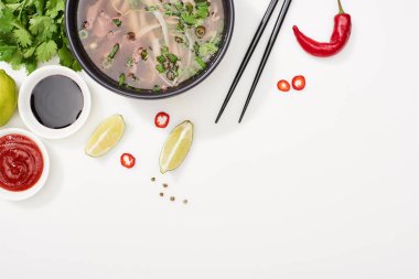 top view of pho in bowl near chopsticks, lime, chili and soy sauces and coriander on white background clipart