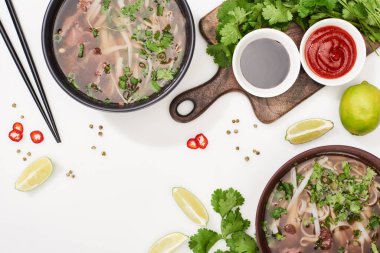 top view of pho in bowls near chopsticks, lime, chili and soy sauces and coriander on white background clipart
