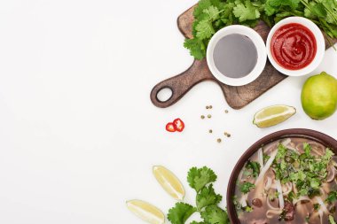 Çubukların, limonun, kırmızı biberin, soya sosunun ve beyaz arka planda kişniş otunun olduğu kasenin en üst görüntüsü.