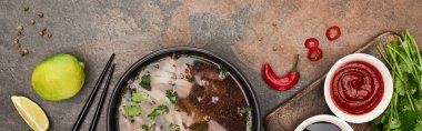 top view of pho in bowl near chopsticks, lime, chili and soy sauces and coriander on stone background, panoramic shot clipart