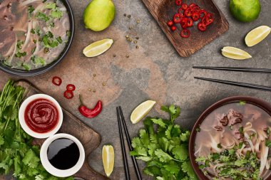 top view of pho in bowls near chopsticks, lime, chili and soy sauces and coriander on stone background clipart