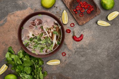 top view of pho in bowl near lime, chili and coriander on stone background clipart