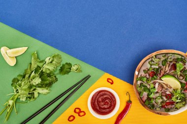 top view of pho in bowl near chopsticks, lime, chili and coriander on blue, yellow, green background clipart