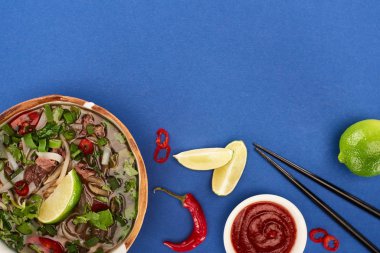 top view of pho in bowl near chopsticks, lime, chili sauce on blue background clipart