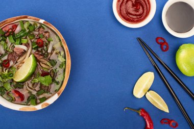 Çubukların, limonun, kırmızı biberin ve mavi arka planda soya sosunun yanındaki kasede pho 'nun üst görüntüsü.
