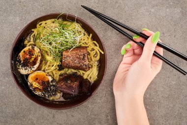 Gri arka planda yumurta ve sığır etli ramen yanında yemek çubuğu tutan bir kadın görüntüsü.