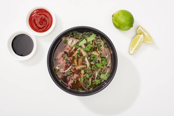 top view of pho in bowl near lime, chili and soy sauces on white background