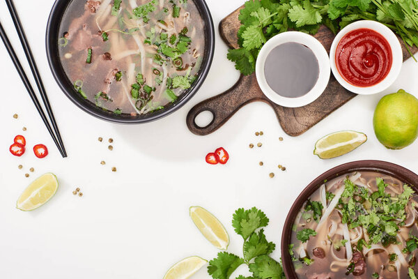 top view of pho in bowls near chopsticks, lime, chili and soy sauces and coriander on white background