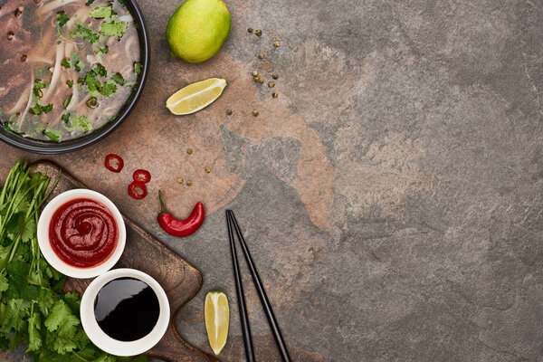 top view of pho in bowl near chopsticks, lime, chili and soy sauces and coriander on stone background