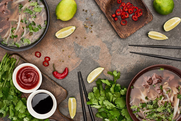 top view of pho in bowls near chopsticks, lime, chili and soy sauces and coriander on stone background