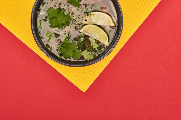 top view of pho in bowl with lime and coriander on red and yellow background