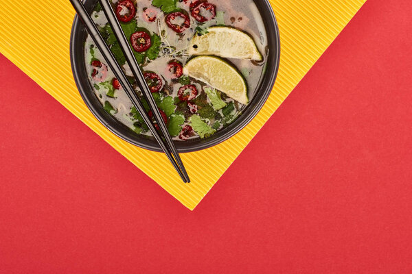 top view of pho in bowl with chopsticks, lime, chili and coriander on red and yellow background