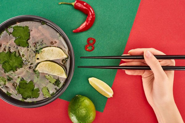 cropped view of woman holding chopsticks near pho in bowl near lime, chili on red and green background