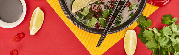 top view of pho in bowl with chopsticks near soy sauce, lime, chili and coriander on red and yellow background, panoramic shot