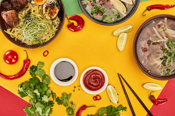 top view of pho and ramen in bowls near lime, chili and soy sauces and coriander on colorful background