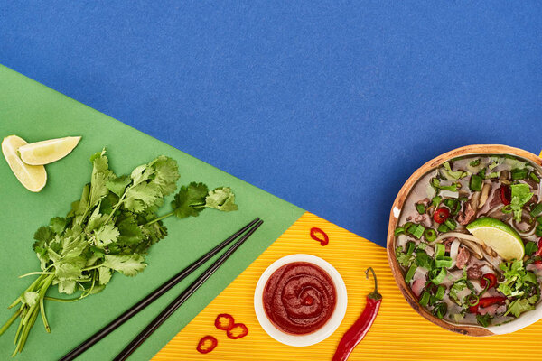 top view of pho in bowl near chopsticks, lime, chili and coriander on blue, yellow, green background