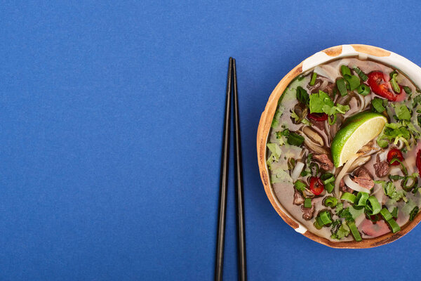 top view of pho in bowl near chopsticks on blue background