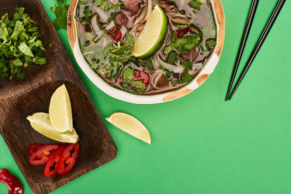 top view of pho in bowl near chopsticks and wooden board with lime, chili and coriander on green background