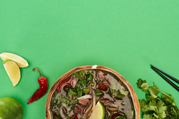 top view of pho in bowl near chopsticks, lime, chili and coriander on green background