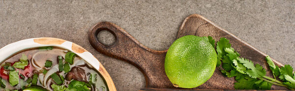 top view of pho in bowl near lime and coriander on wooden cutting board on grey background, panoramic shot