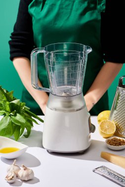 Pesto sosunun yanında duran kadının kırpılmış görüntüsü ham malzemeler ve yeşil masada izole edilmiş yemek işlemcisi.