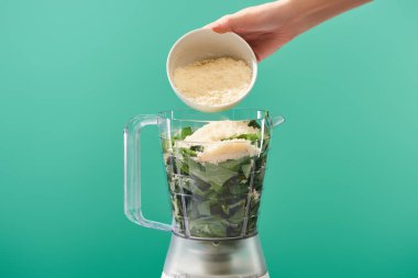 cropped view of woman adding grated Parmesan to basil leaves in food processor isolated on green clipart