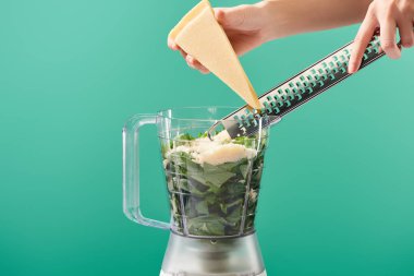 cropped view of woman grating Parmesan in food processor with basil leaves isolated on green clipart