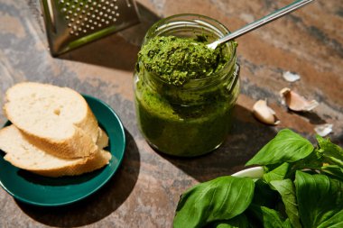 baget dilimleri, kavanozda pesto sosu, rende ve taş yüzeyinde taze malzemeler.