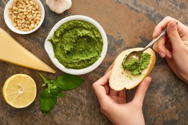 Kırpılmış bir kadın, ekmek dilimine pesto sosu ekliyor. Taş yüzeyindeki taze malzemelerin yanında.