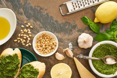 Pesto soslu baget dilimlerinin üst görüntüsü taze malzemelerin yanında ve taş yüzeyde pişirme aletleri.