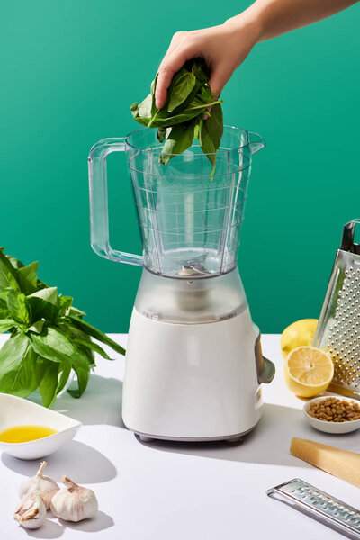 cropped view of woman putting basil leaves in food processor near pesto sauce raw ingredients on white table isolated on green