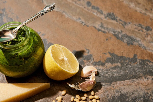 pesto sauce in glass jar with spoon near ingredients on stone surface
