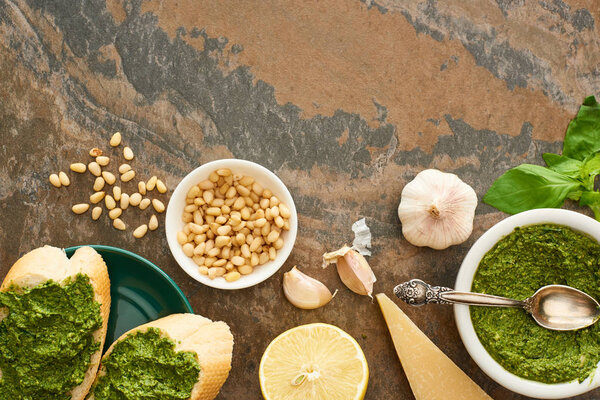 top view of baguette slices with pesto sauce on plate near fresh ingredients on stone surface