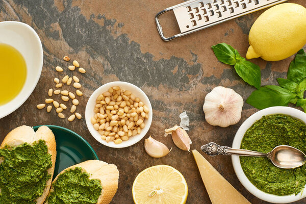top view of baguette slices with pesto sauce on plate near fresh ingredients and cooking utensils on stone surface
