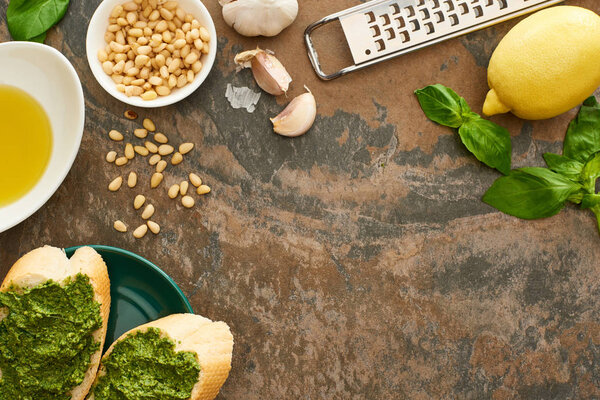top view of baguette slices with pesto sauce on plate near fresh ingredients and cooking utensils on stone surface