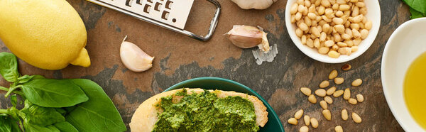 top view of baguette slice with pesto sauce on plate near fresh ingredients and cooking utensils on stone surface, panoramic shot
